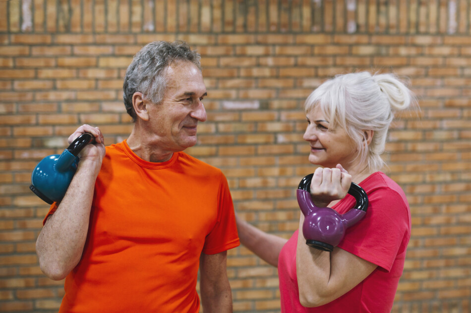 fotografía de dos adultos mayores sonriéndose entre si mientras realizan ejercicios de pesas en un club deportivo