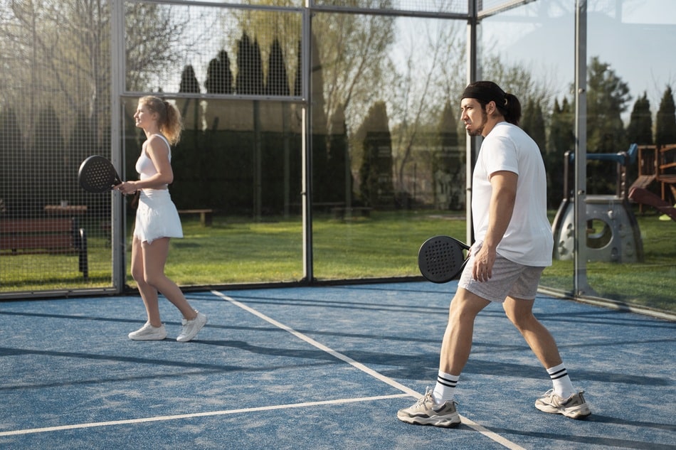 hombre y mujer disputando un partido de padel outdoor a plena luz del dia con arboles y juegos infantiles en segundo plano